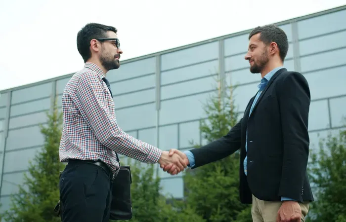 Two business professionals shaking hands outside modern office building surrounded by green trees, symbolizing partnership